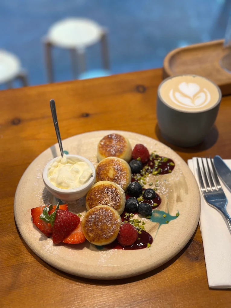 Assiette de syrniki dorés, crème et fruits rouges, à côté d'un flat white au latte art sur une table en bois, chez Sublime Coffee à Genève.